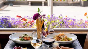 A dining table with various dishes and glasses of wine, set against a backdrop of a garden with flowers and a view of the sea.