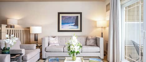 Una sala de estar moderna con un sofá gris, dos sillones, una mesa de centro de vidrio con libros y un jarrón de flores, un cuadro en la pared y un amplio ventanal con cortinas.