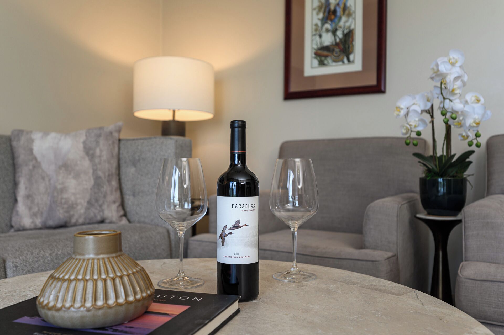 A table with a bottle of wine, two wine glasses, a book, and a decorative vase.