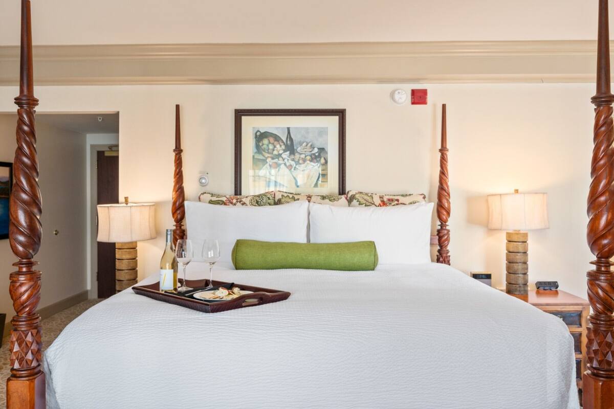 A four-poster bed with a tray of food and wine, a green pillow, and a framed artwork above.