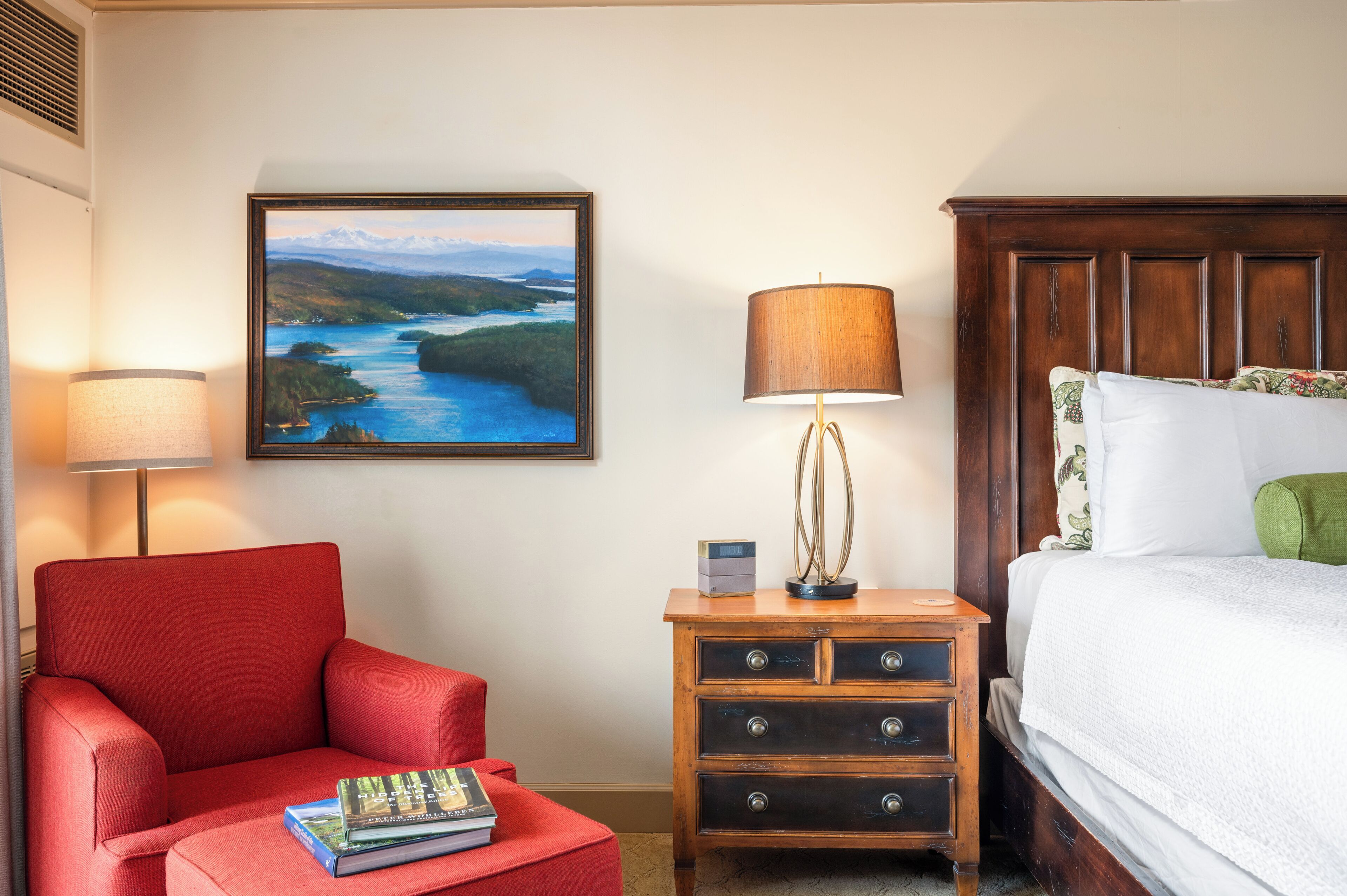 A hotel room with a bed, a red armchair, a wooden nightstand with a lamp, and a framed landscape painting on the wall.