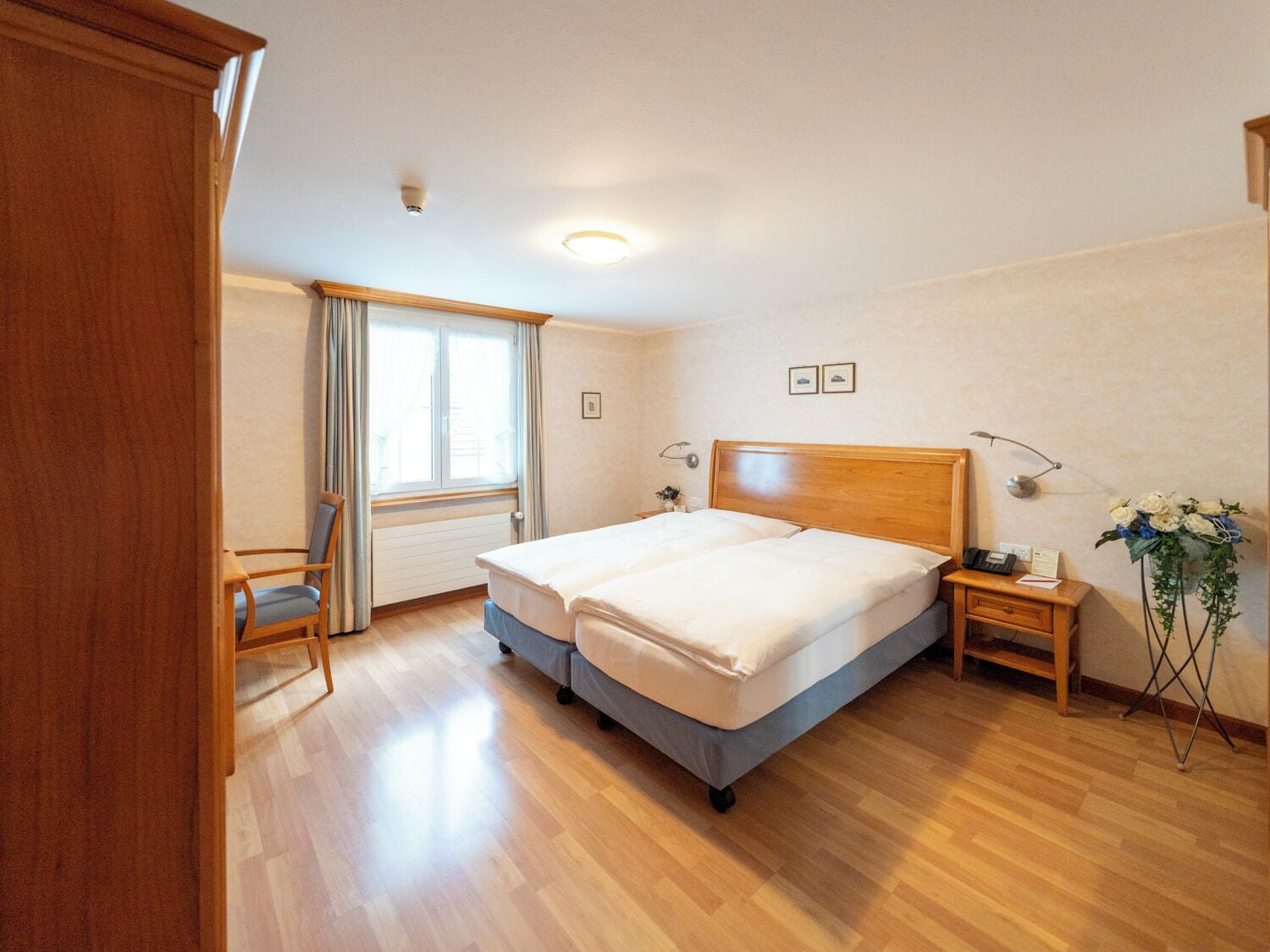 A double bed with a wooden headboard, a bedside table with a lamp, a chair, and a window with curtains.