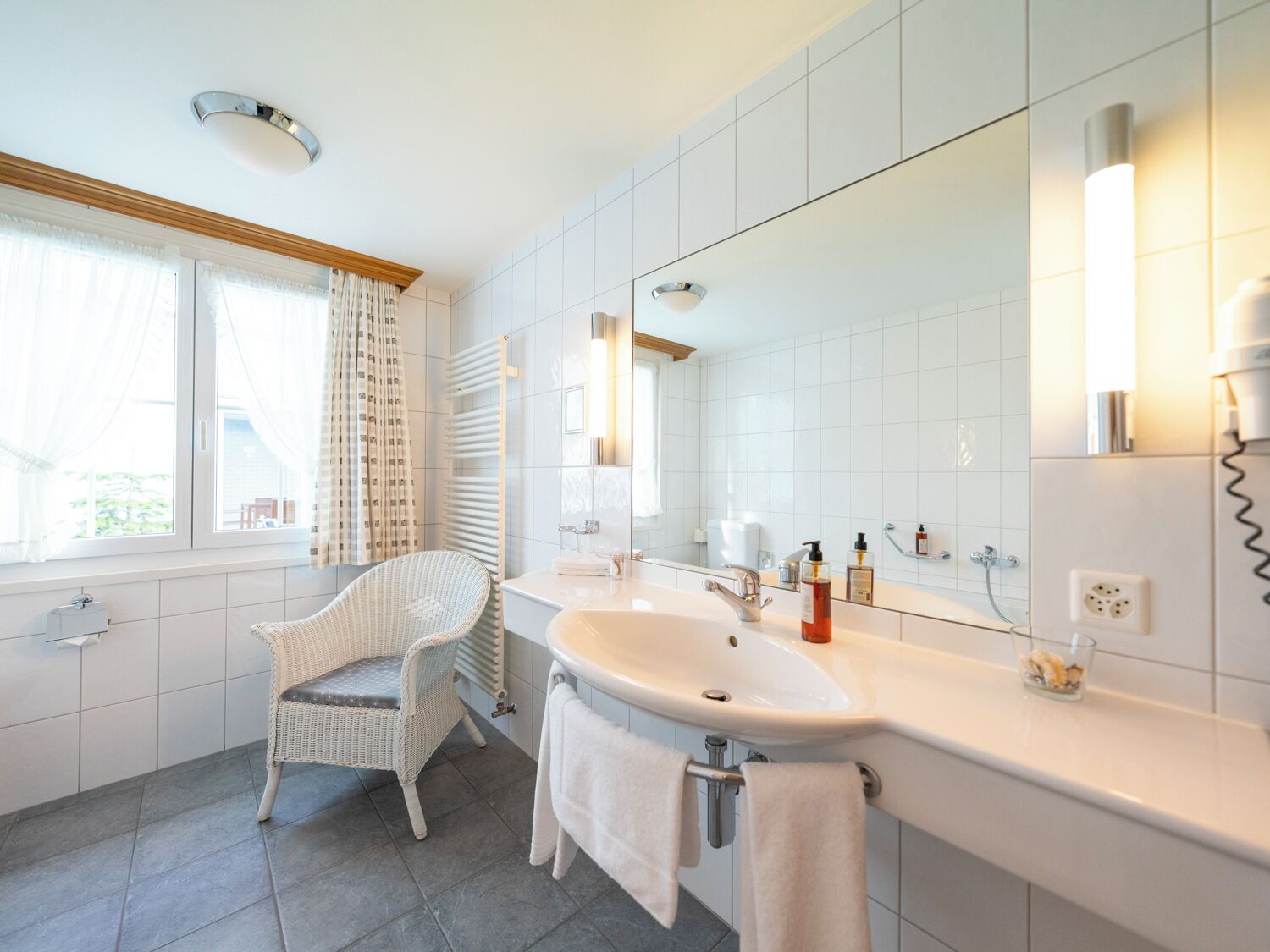 A modern bathroom with a large mirror, white tiles, a wicker chair, and a bathtub.
