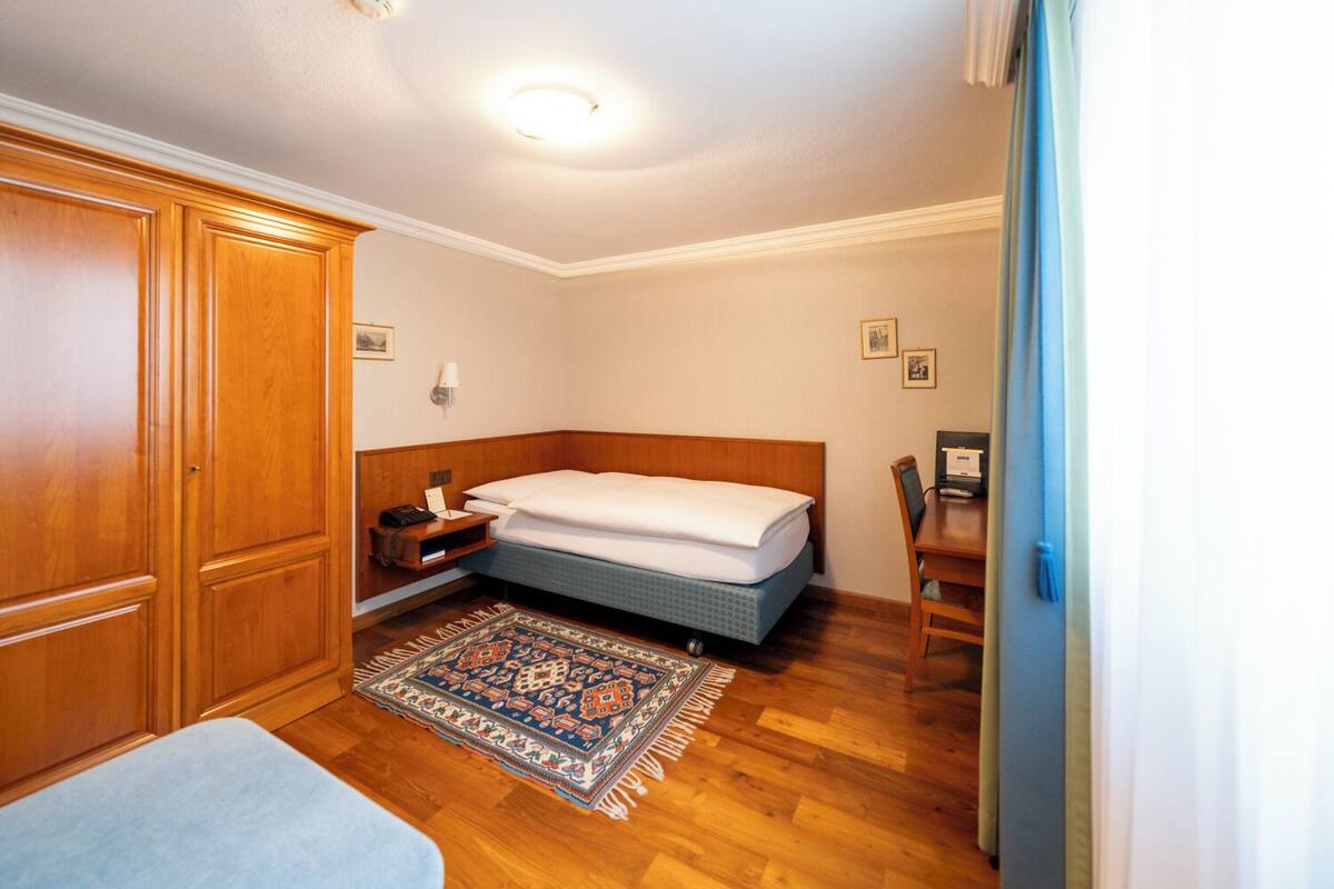 Habitació d'hotel amb un armari de fusta, un llit amb capçalera blava, una taula de nit amb una làmpada i una catifa petita a terra.