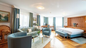 A modern hotel room with a sofa, armchair, and a bed. There is a desk with a chair, a lamp, and a vase with flowers.