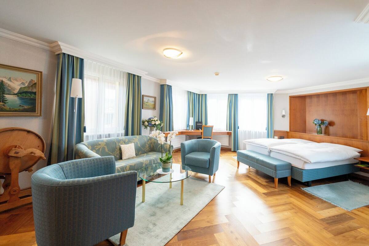 Habitació d'hotel moderna amb un sofà, una butaca i un llit. Hi ha una taula de treball amb una cadira, una làmpada i un vas amb flors.