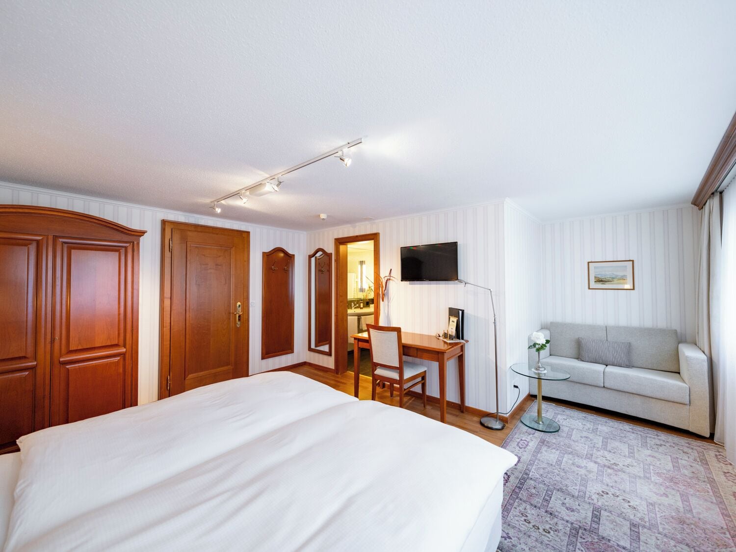 A hotel room with a large bed, a desk with a chair, a television, a sofa, and a small table with a lamp.