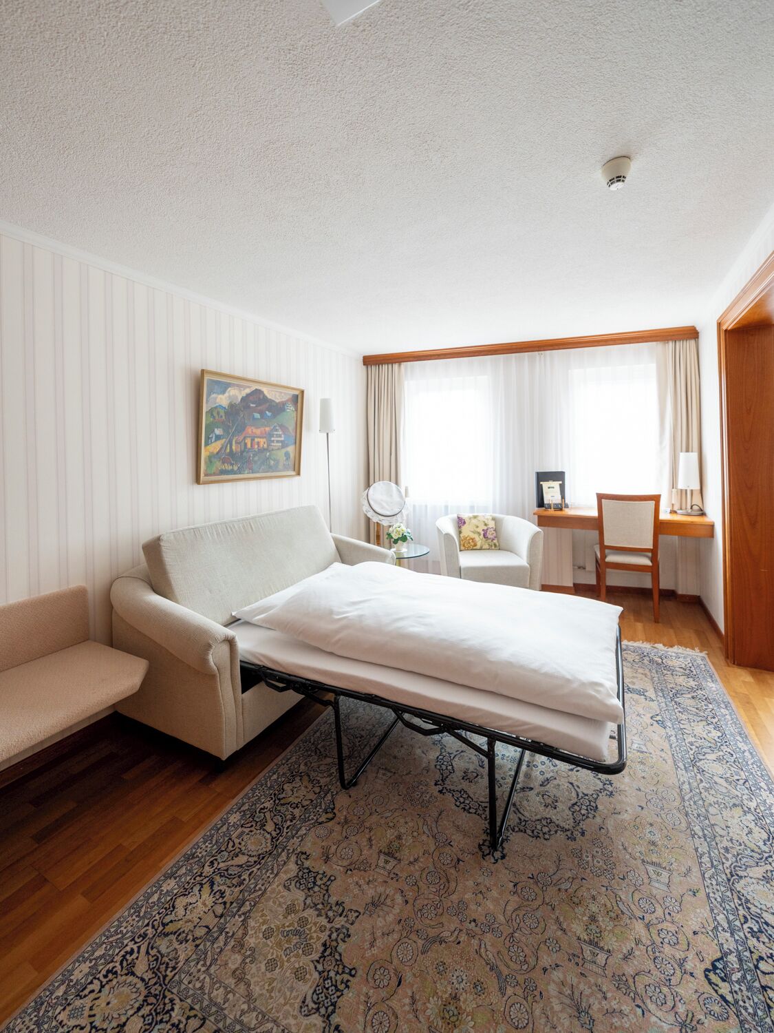 A hotel room with a bed, a desk, a chair, a small table, and a painting on the wall.
