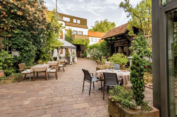 Un patio al aire libre con mesas y sillas, rodeado de follaje y edificios.