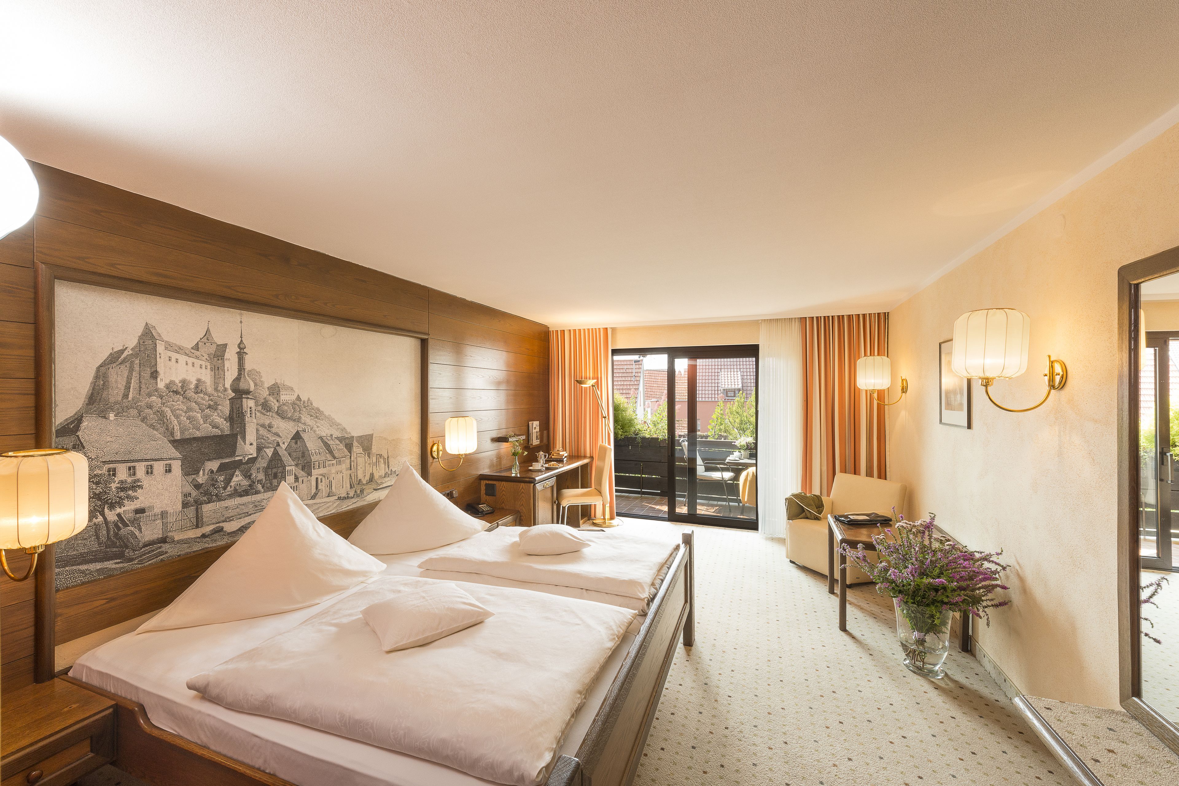 Habitación de hotel con dos camas, un escritorio, una silla, un florero con flores y un mural en la pared.