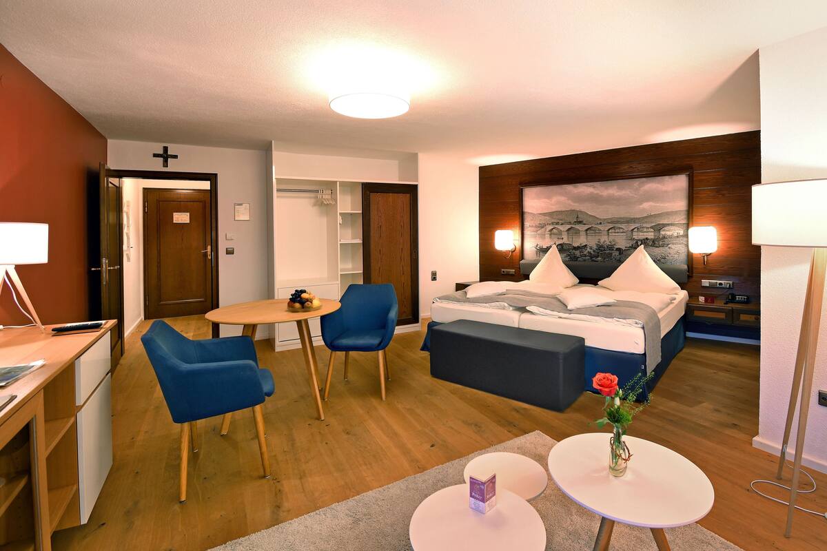 Quarto de hotel com cama, escrivaninha, duas cadeiras, uma mesinha e um quadro na parede.