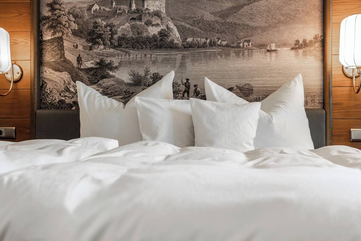 Quarto de hotel com cama, almofadas brancas e um grande mural na parede retratando um castelo à beira de um lago.