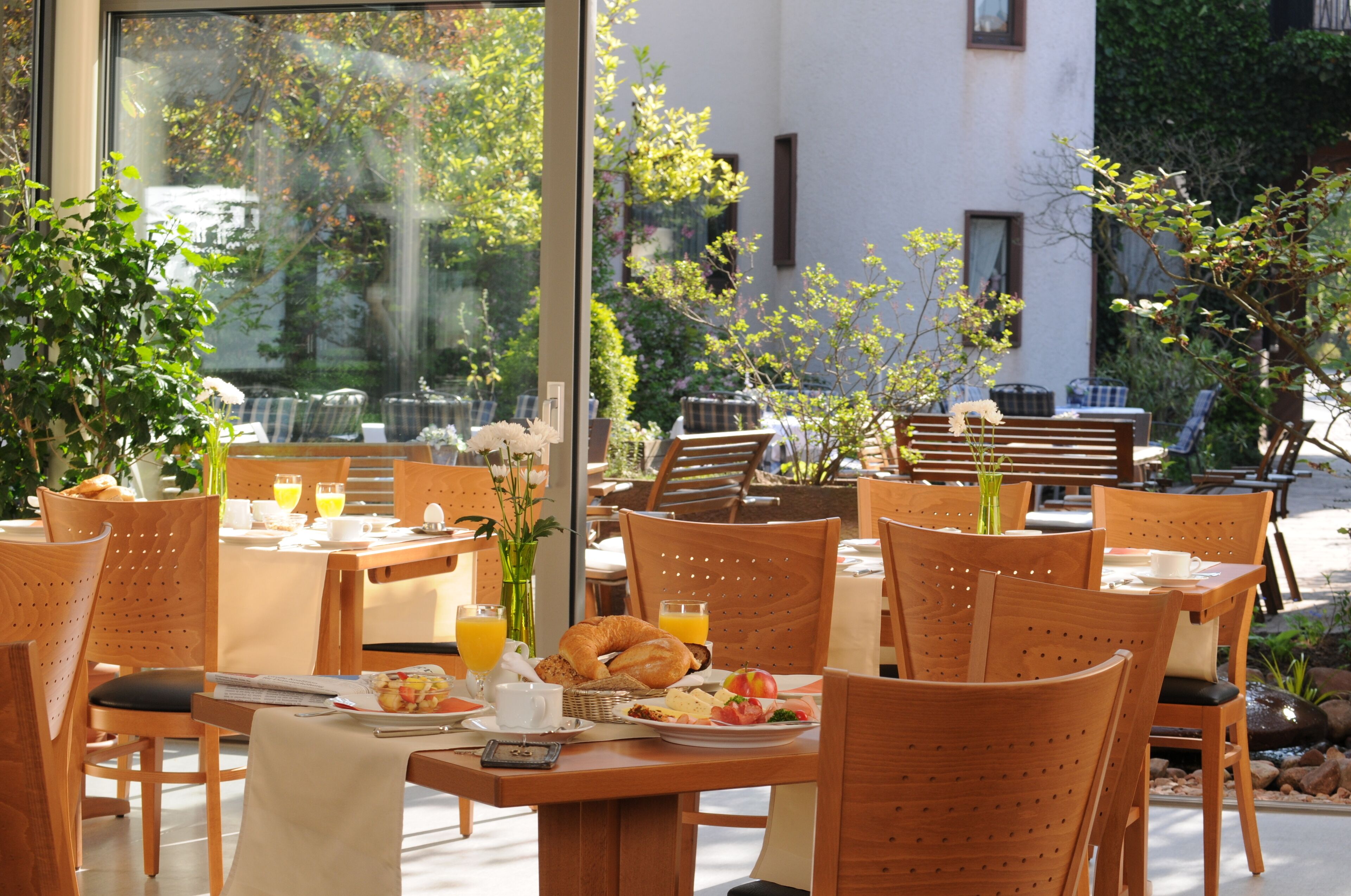 Un área de comedor al aire libre con mesas y sillas de madera, alimentos para el desayuno y vistas al follaje circundante.
