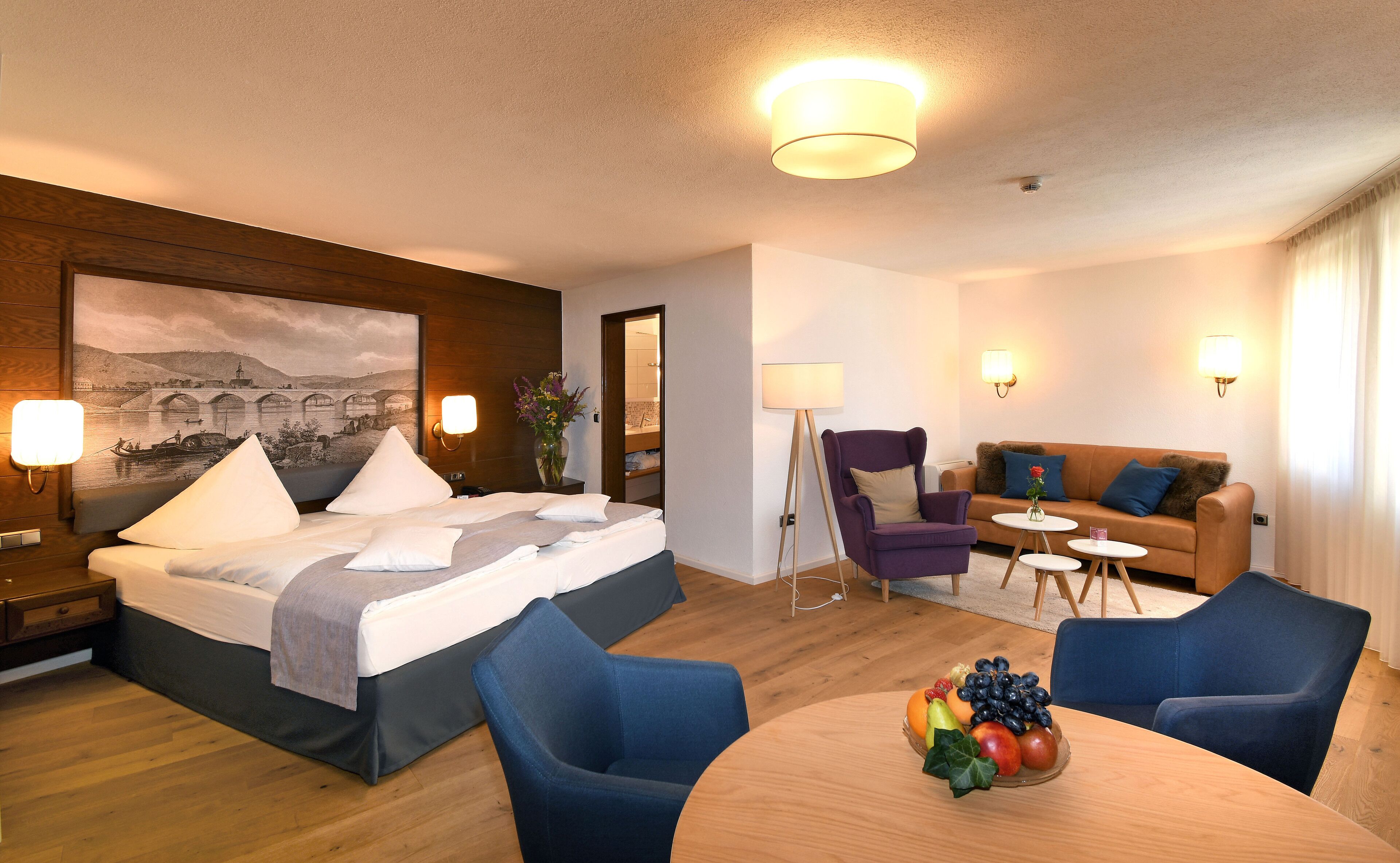 Une chambre d’hôtel comprenant un lit, un bureau, une chaise, un canapé et une table à manger avec des fruits.