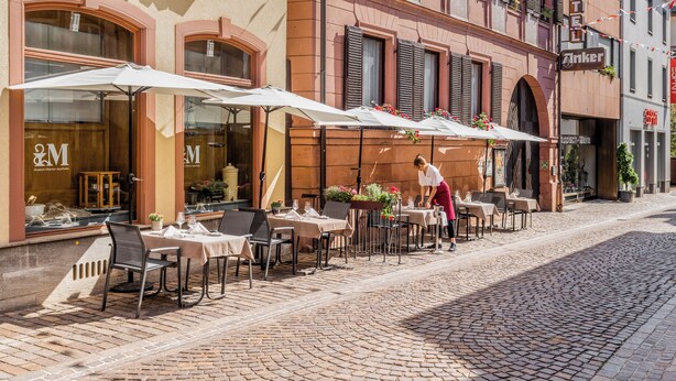 An outdoor café with tables and chairs, a waiter, and a building with a sign that reads "Hotel Anker". - Hotel Anker (Marktheidenfeld)