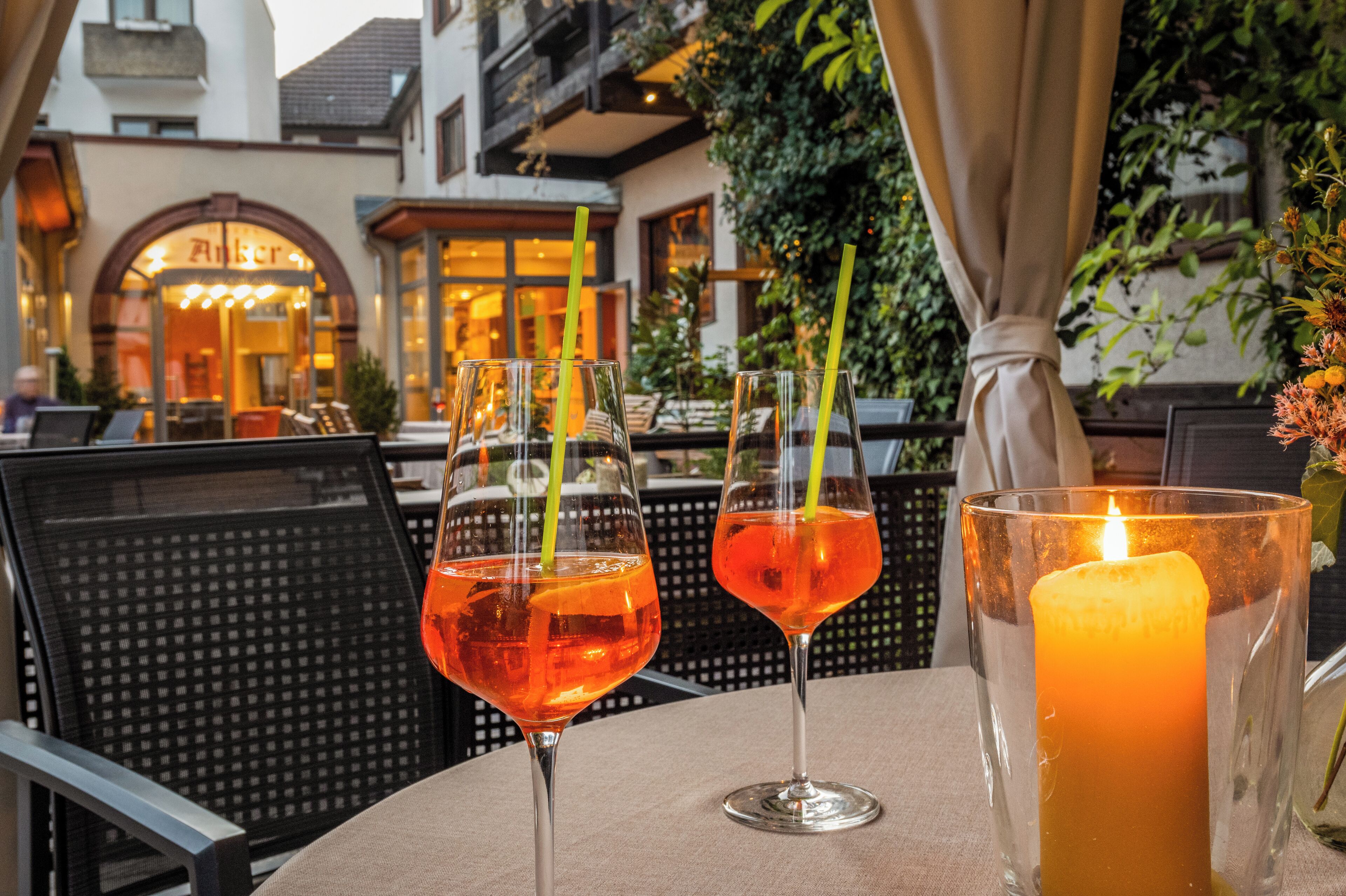 Une table avec deux boissons et une bougie, placée devant un bâtiment dont l’entrée est en arc de cercle et qui est entouré de verdure.