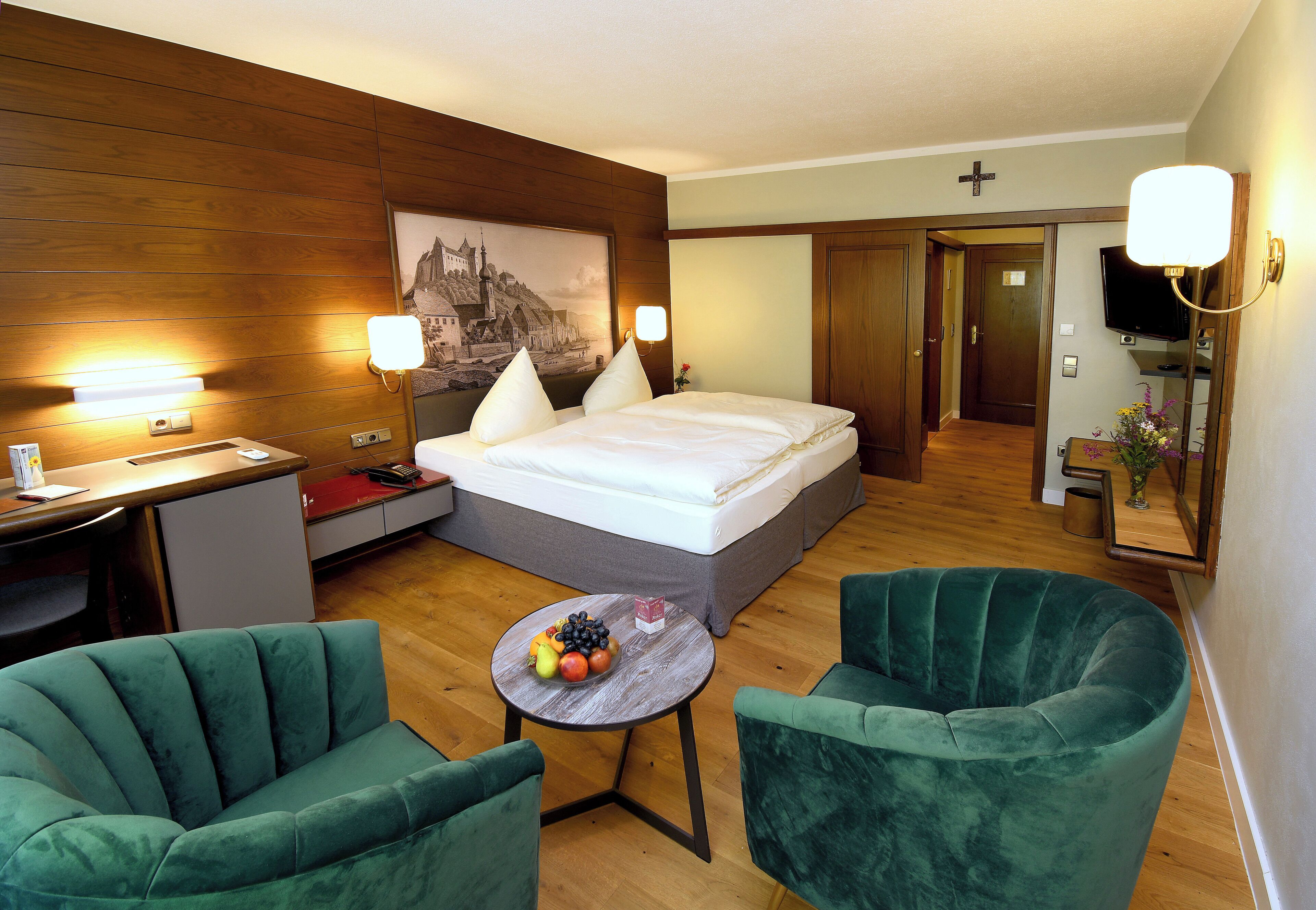 A hotel room with a bed, two green armchairs, a small table with fruit, a desk, and a TV.