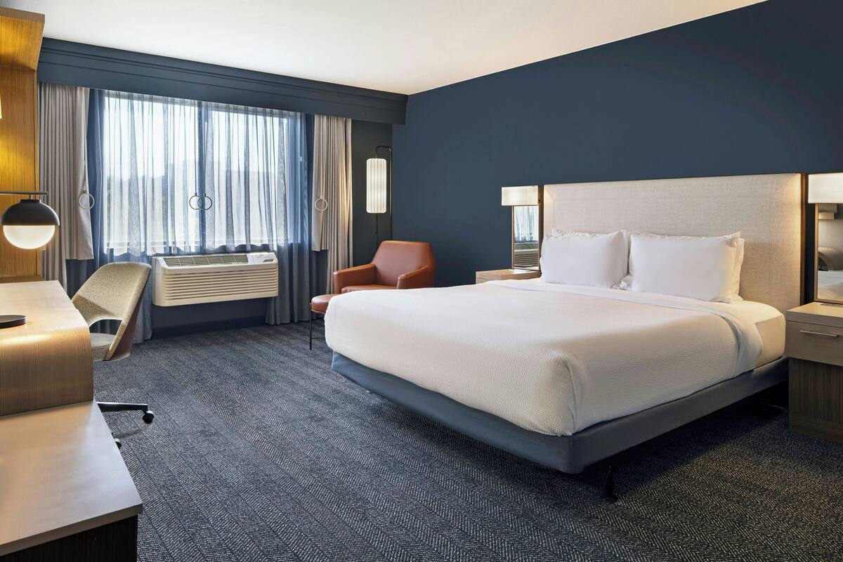A hotel room with a large bed, a desk, a chair, and an air conditioning unit.