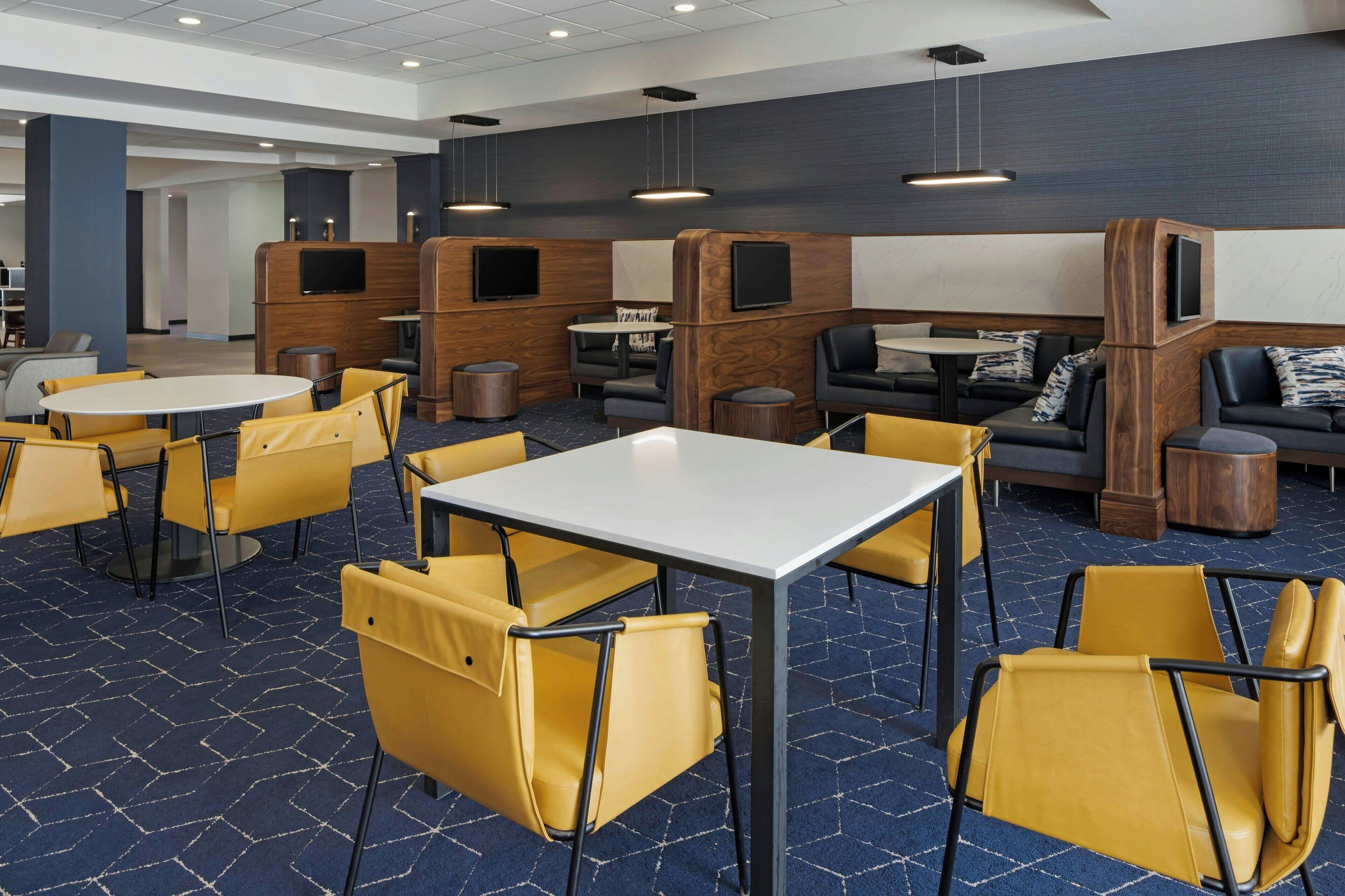A modern hotel lobby with yellow chairs and tables, wooden partitions, and a flat-screen TV mounted on the wall.