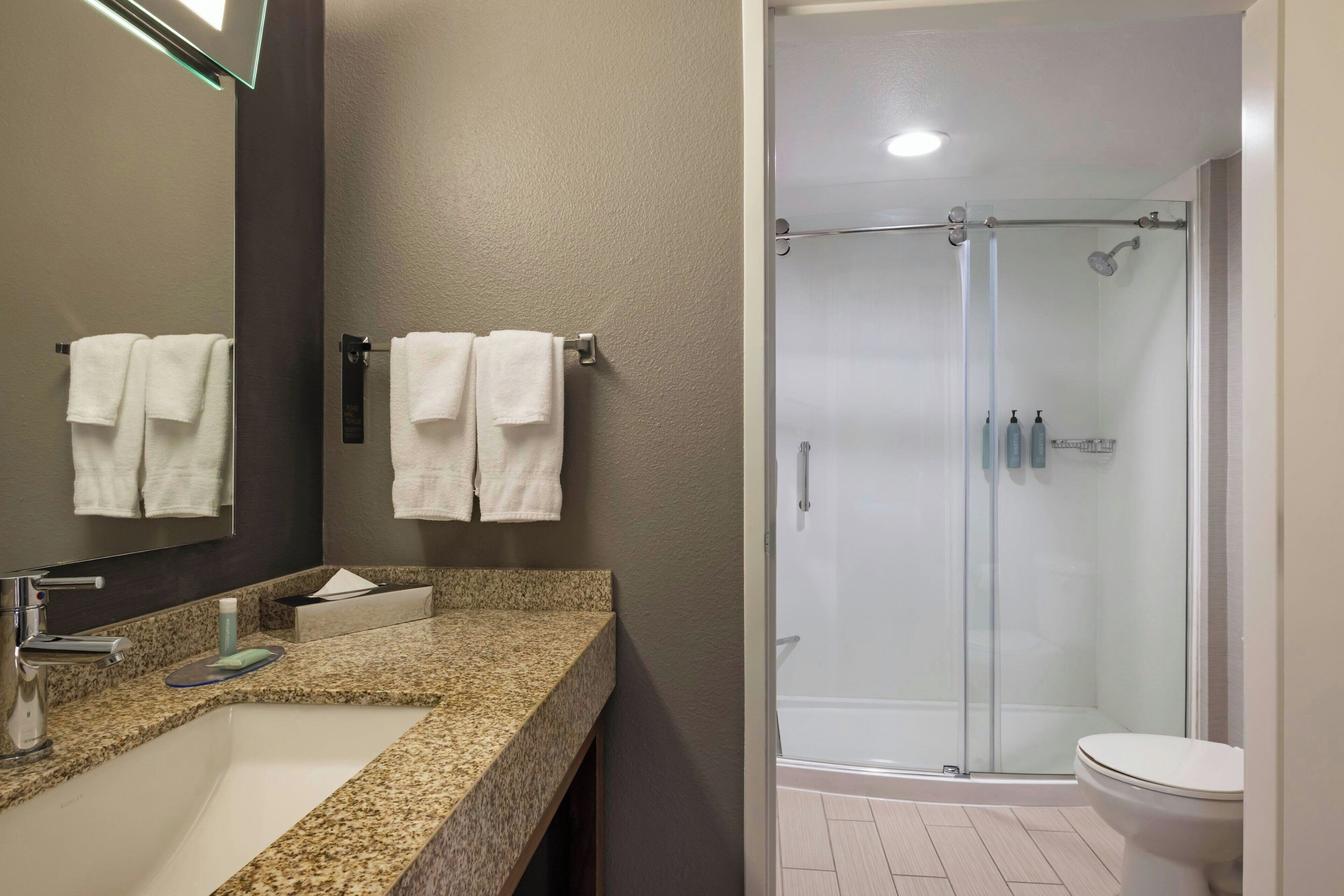A bathroom with a granite countertop sink, a mirror, two white towels, a glass shower enclosure, and a toilet.