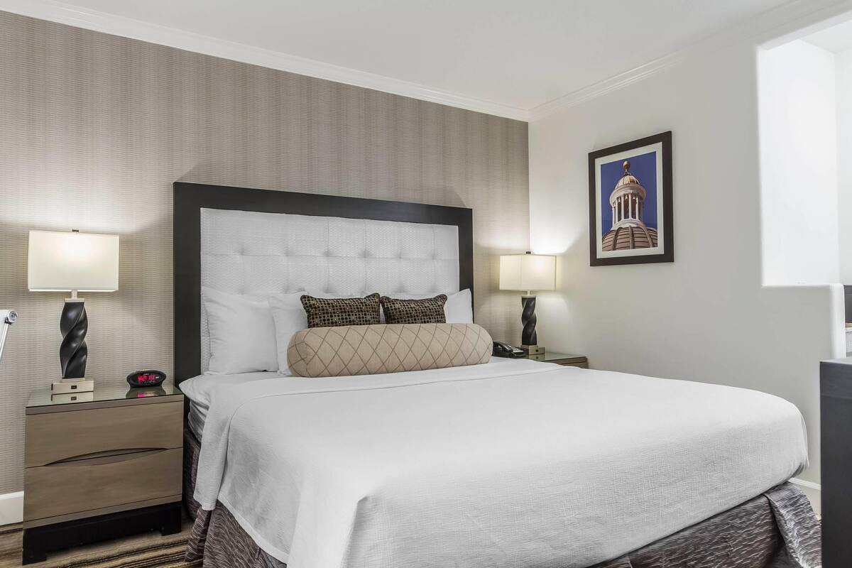 Une chambre d’hôtel avec un grand lit, deux tables de chevet, une petite table, une lampe et un tableau encadré au mur.
