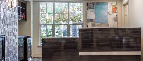 Un espace d’accueil moderne, doté d’un comptoir de réception en bois foncé, d’une cloison en verre et d’une grande fenêtre offrant une vue sur les arbres et les bâtiments.