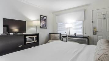 A hotel room with a bed, a TV, a desk with a lamp, and a window with blinds.
