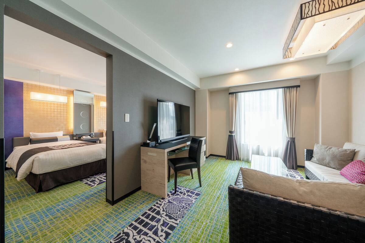 A hotel room with a bed, a desk with a television, a sofa, and a large window with curtains.