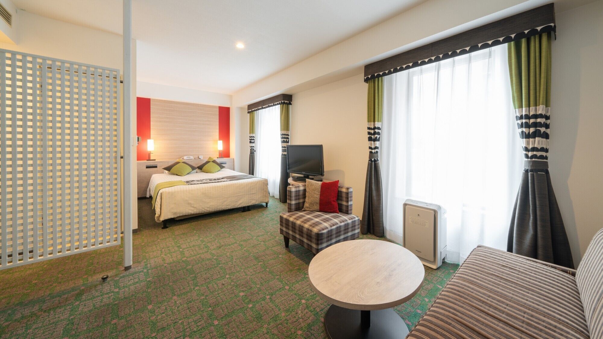A hotel room with a bed, a TV, a small table, a sofa, and a window with curtains.