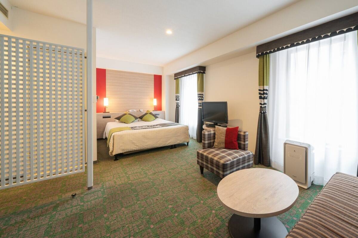 A hotel room with a bed, a TV, a small table, a sofa, and a window with curtains.