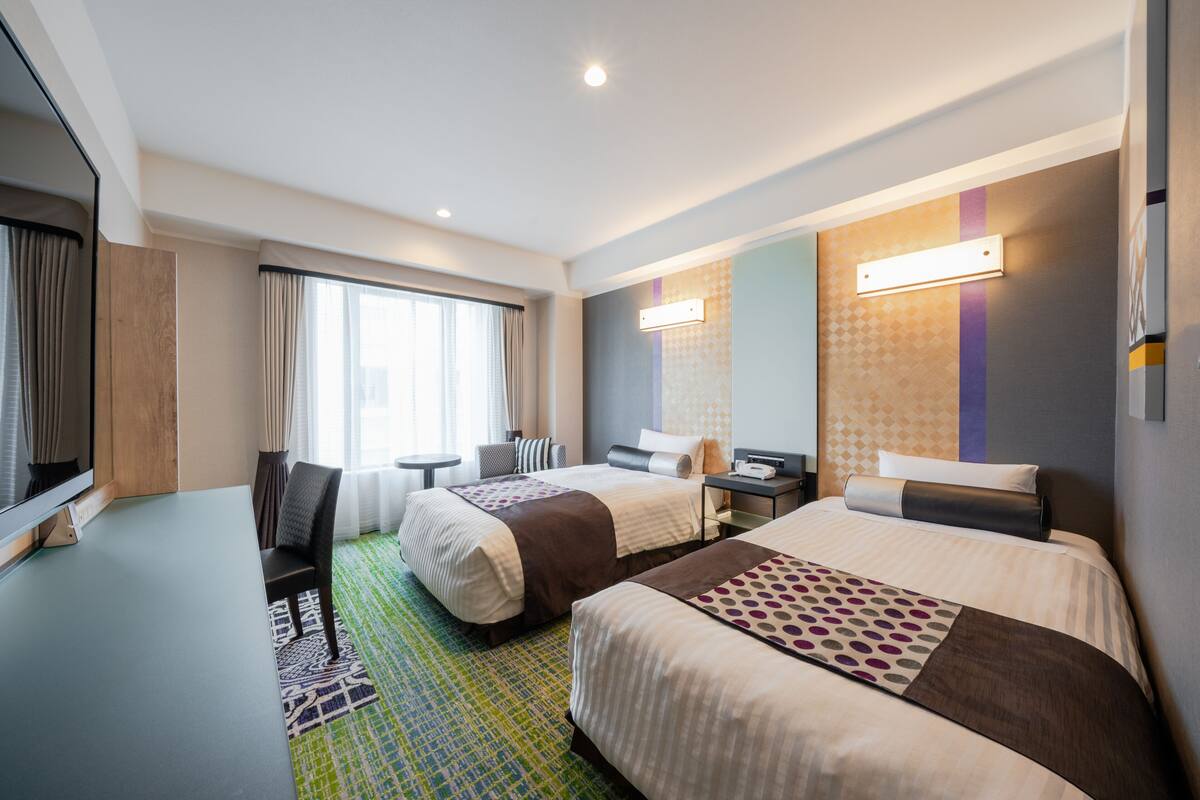A hotel room with two beds, a desk, a chair, a TV, and a window with curtains.