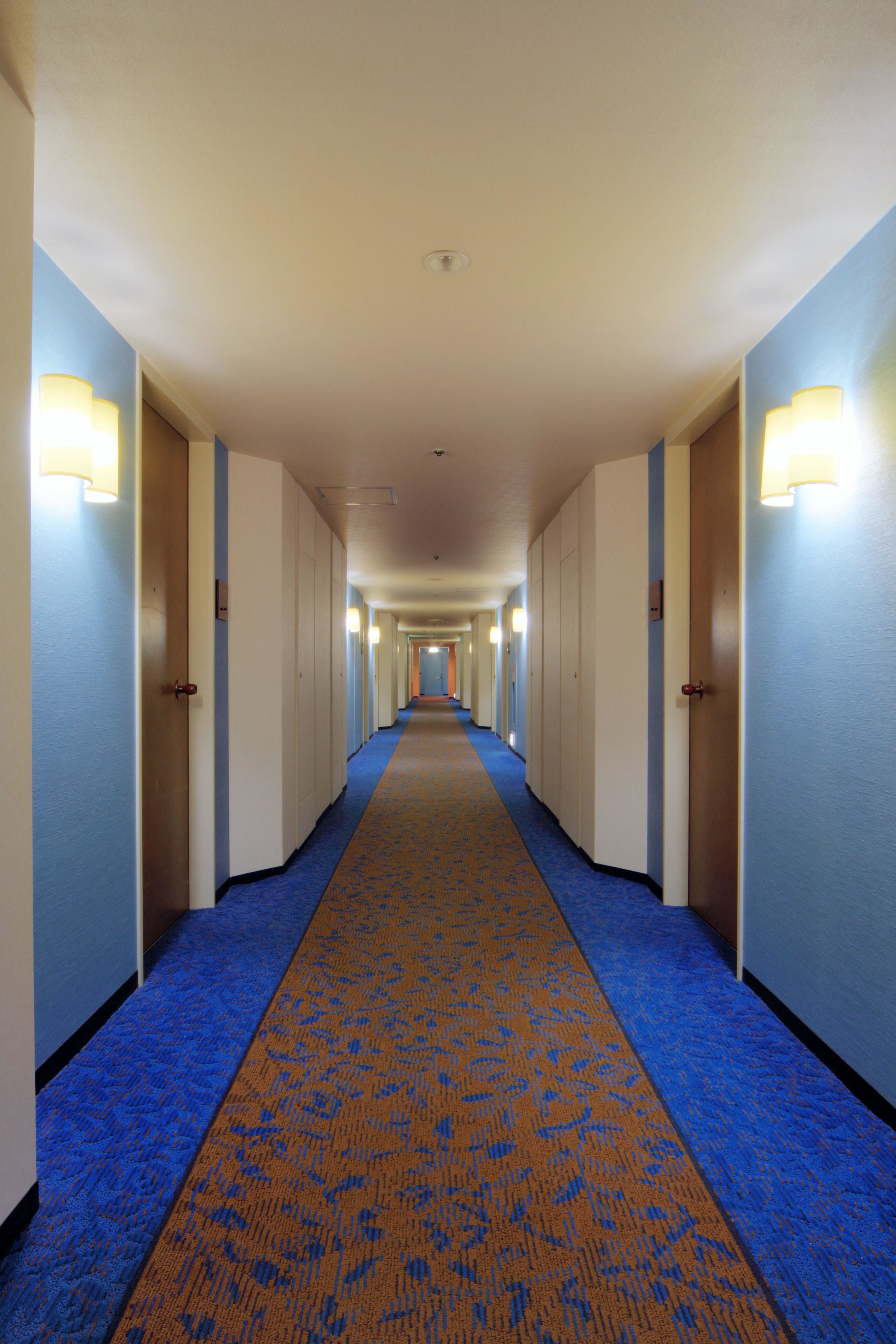 A hotel corridor with a blue carpet, doors on both sides, and wall-mounted lights.