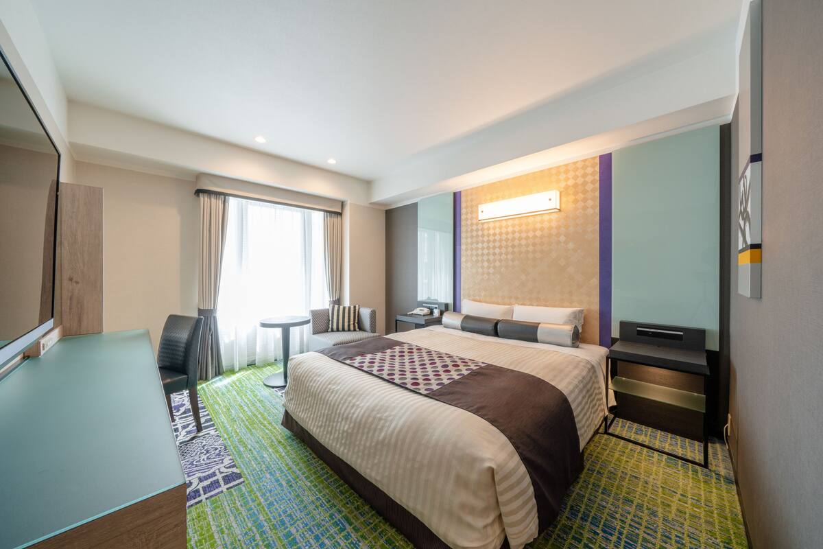A hotel room with a bed, a desk, a chair, a small table, and a TV.