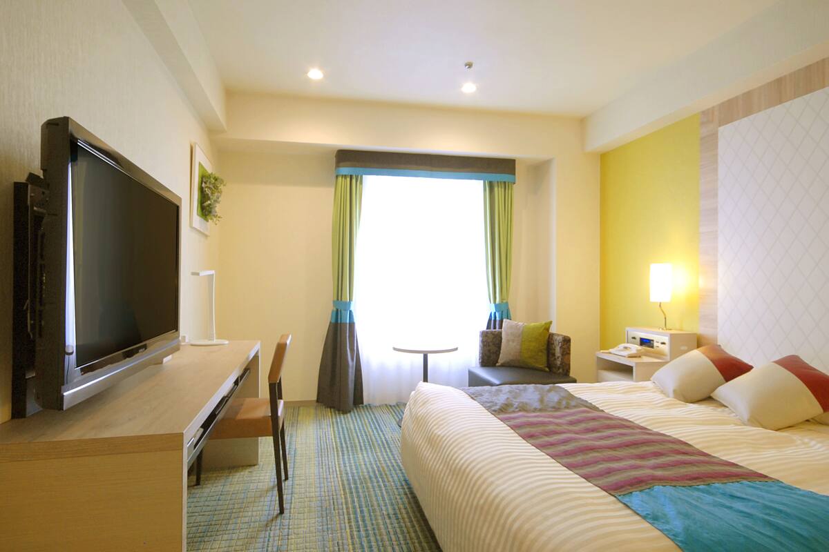 A hotel room with a bed, a desk with a flat-screen TV, a small table, and a lamp.