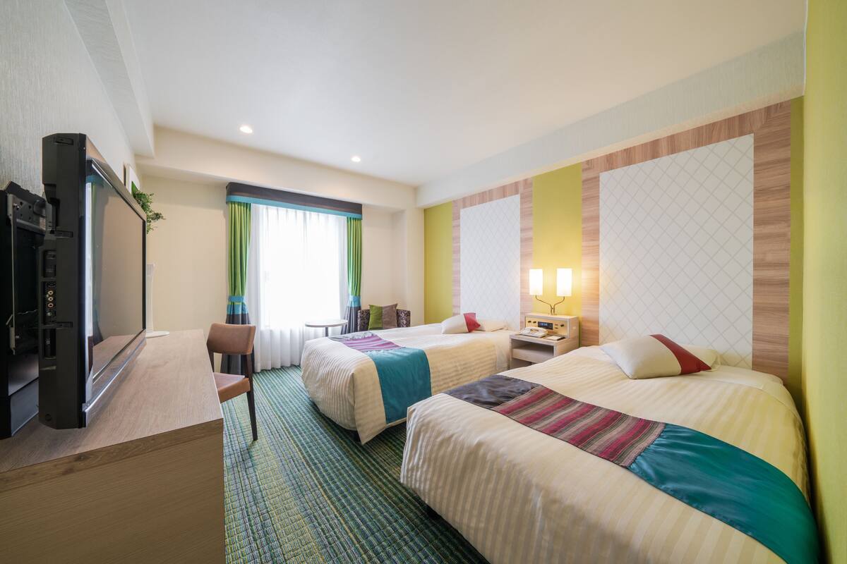 A hotel room with two beds, a desk, a TV, and a window with curtains.