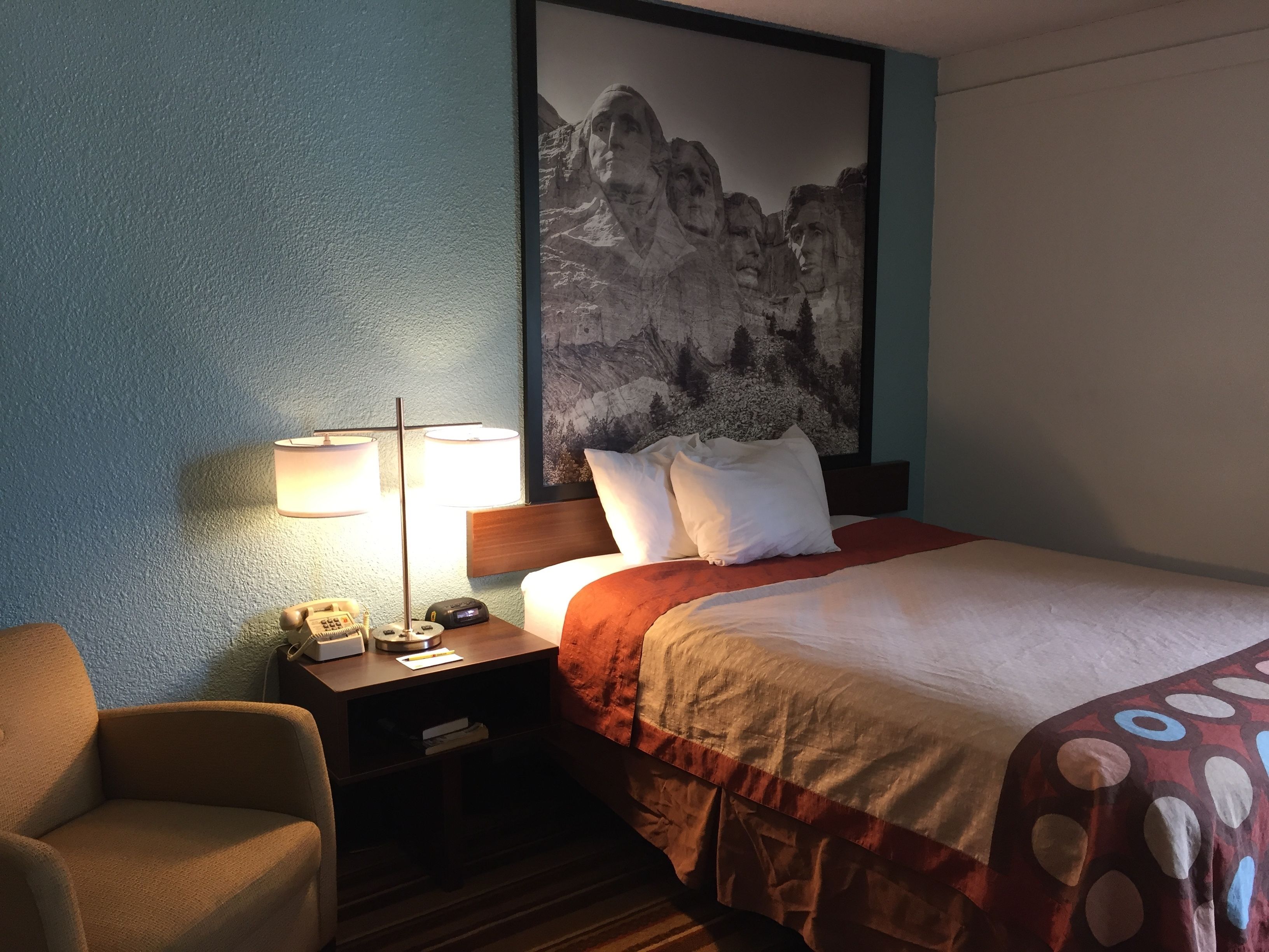 Quarto de hotel com cama, mesa de cabeceira com abajur, uma cadeira e um quadro na parede.