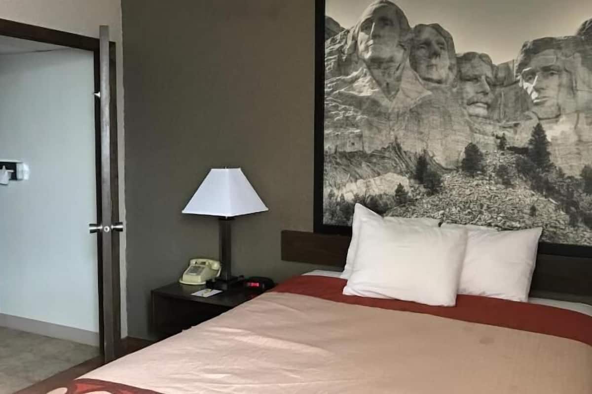Een hotelkamer met een bed, een nachtkastje met een lamp, een telefoon en een groot schilderij van een berglandschap aan de muur.