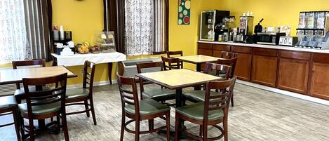 Un comedor con mesas y sillas de madera, un reloj en la pared y una zona de cocina con electrodomésticos.