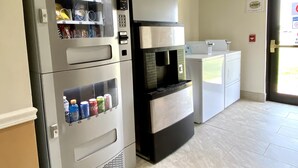 A vending machine with snacks and drinks, a coffee machine, and a washing machine in a room with a tiled floor and a door with an exit sign. - FairBridge Inn Express Mount Vernon (Mount Vernon)