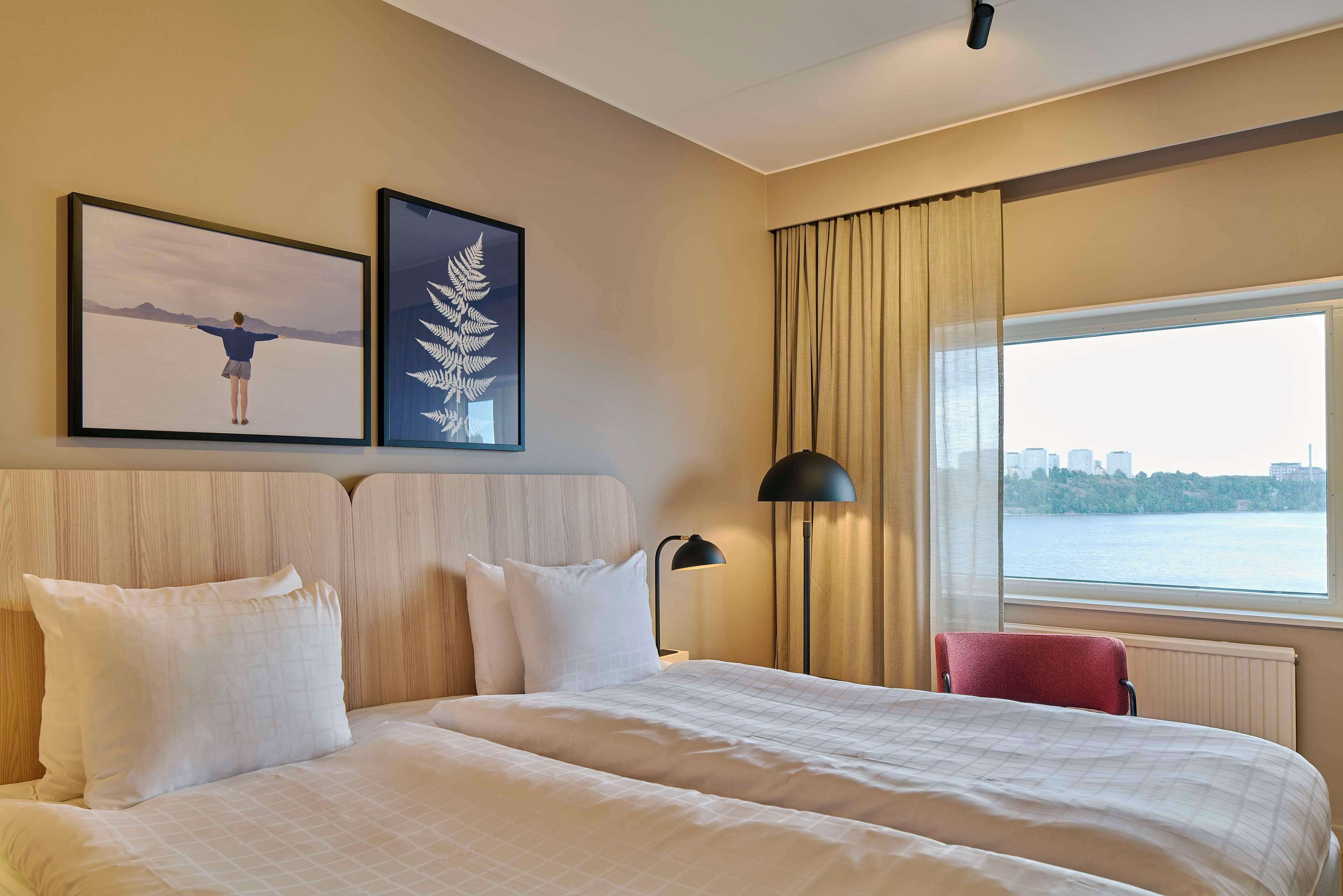 A hotel room with a large bed, two pillows, a nightstand with a lamp, a red chair, and a window with a view of a city skyline.