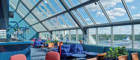 A sunlit deck with seating, a bar, and a view of a cruise ship.