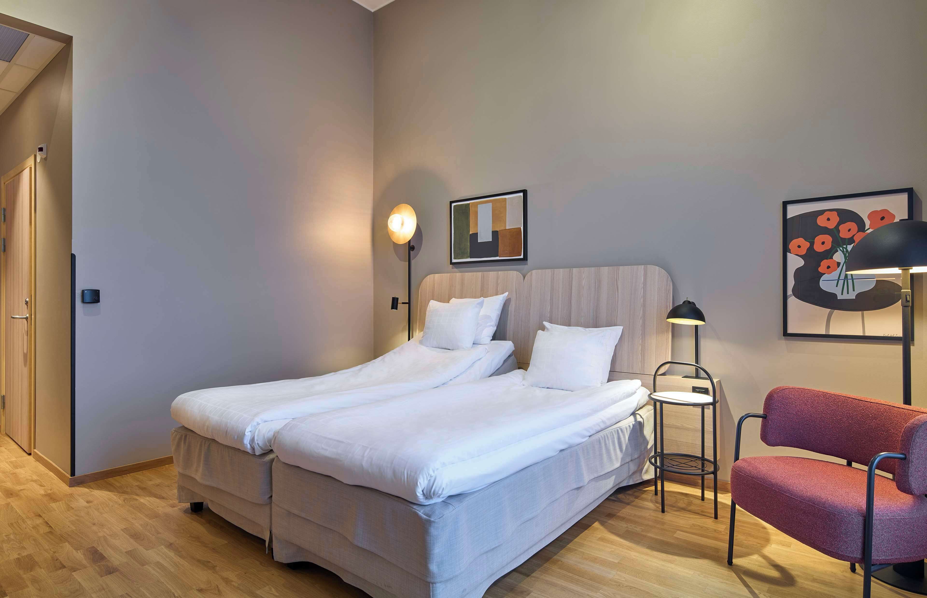 Quarto de hotel moderno com uma cama grande, uma mesinha de apoio, uma cadeira vermelha e obras de arte nas paredes.