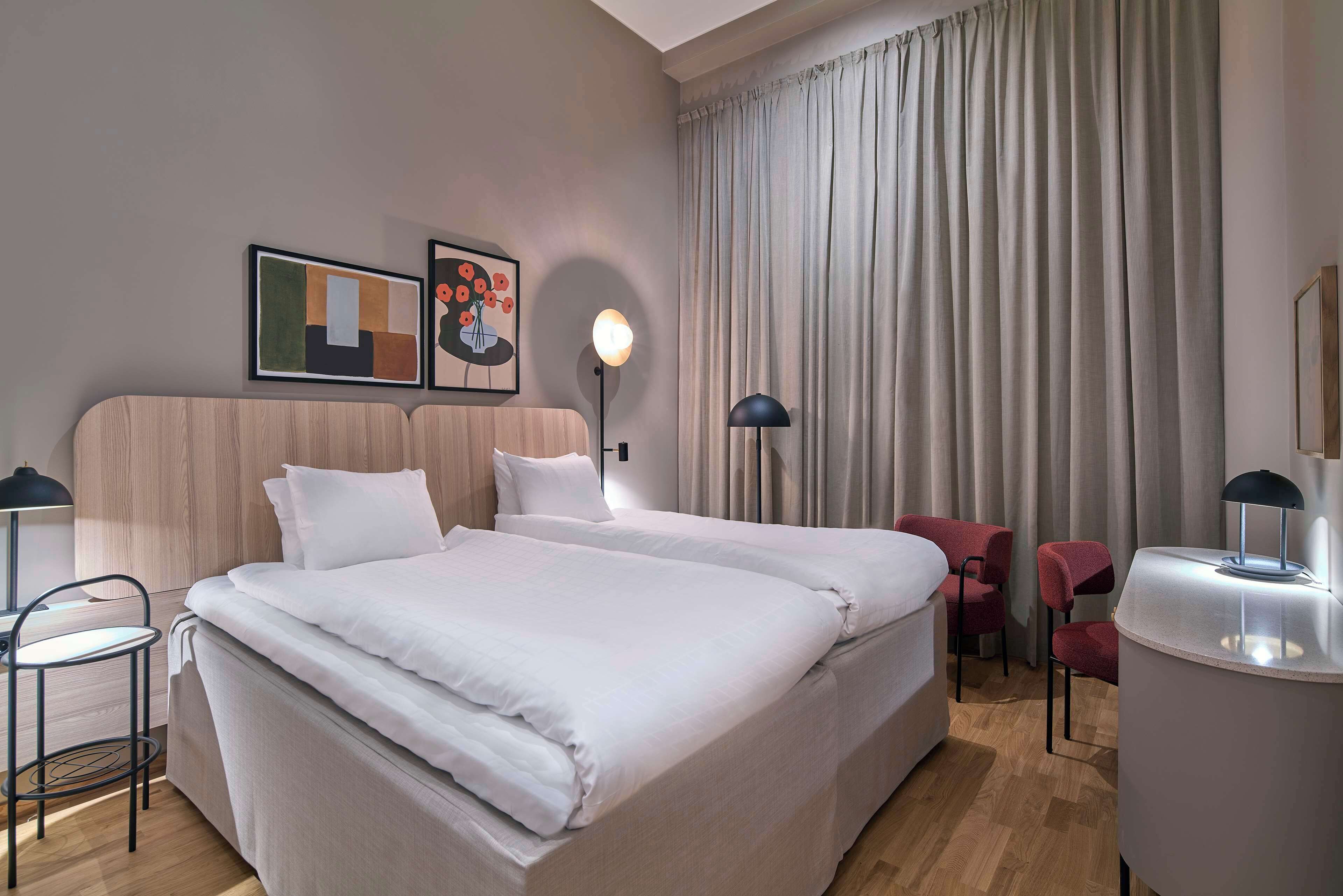 Habitación de hotel con una cama grande, dos mesitas de noche, un escritorio y una silla. Hay dos obras de arte enmarcadas en la pared y un ventanal con cortinas.