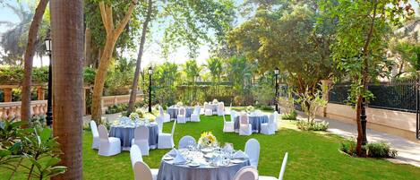 An outdoor dining area with round tables set for a formal event, surrounded by white chairs and greenery.