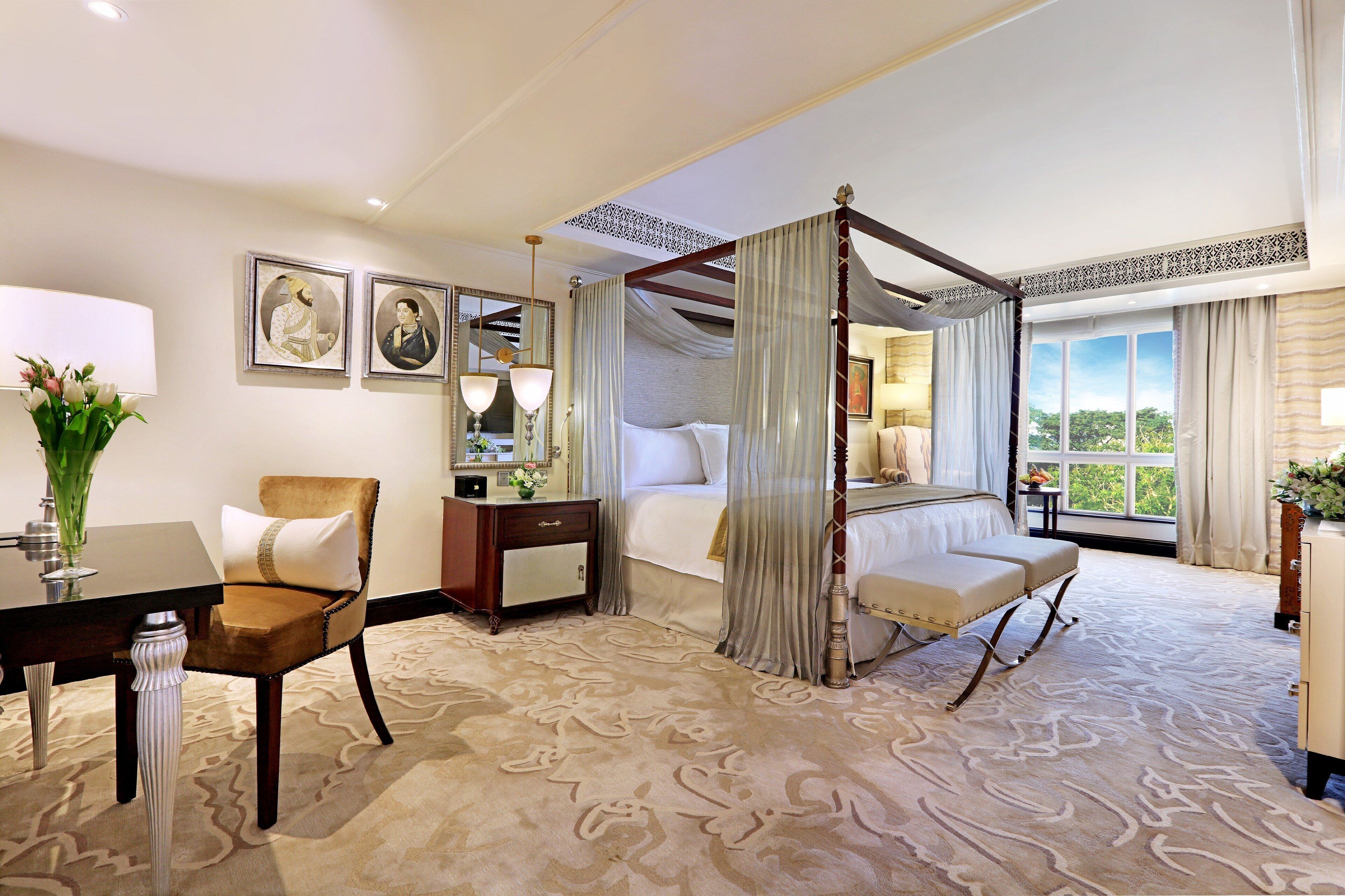 A hotel room with a four-poster bed, a sitting area with a bench, a dining table with a chair, and a large window offering a view.
