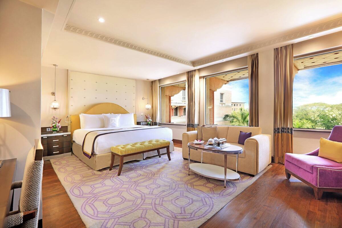 Ein Hotelzimmer mit einem großen Bett, einem Schlafsofa, einem kleinen Tisch und einem Sessel. Ein Fenster bietet Blick auf Gebäude und Bäume.