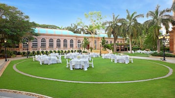 Un patio con varias mesas redondas dispuestas para comer, rodeado de un césped bien cuidado y palmeras.