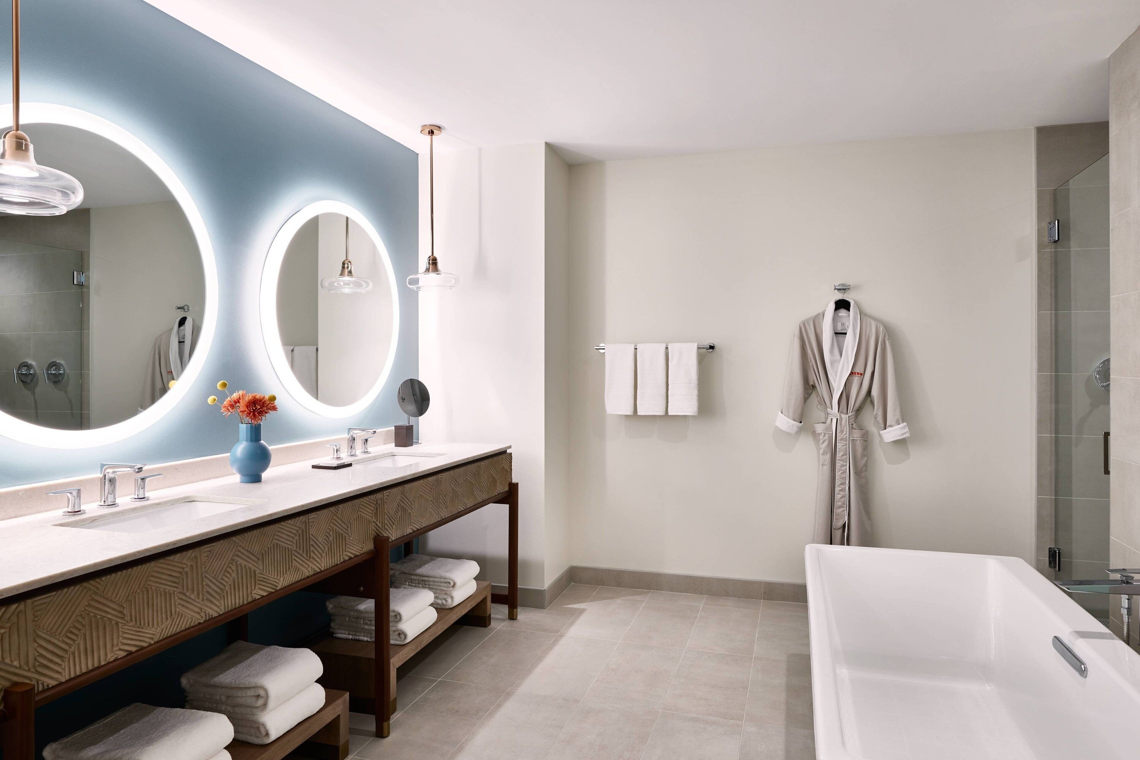 A modern bathroom with a large mirror, two sinks, a vanity with shelves, a bathtub, and a robe hanging on the wall.