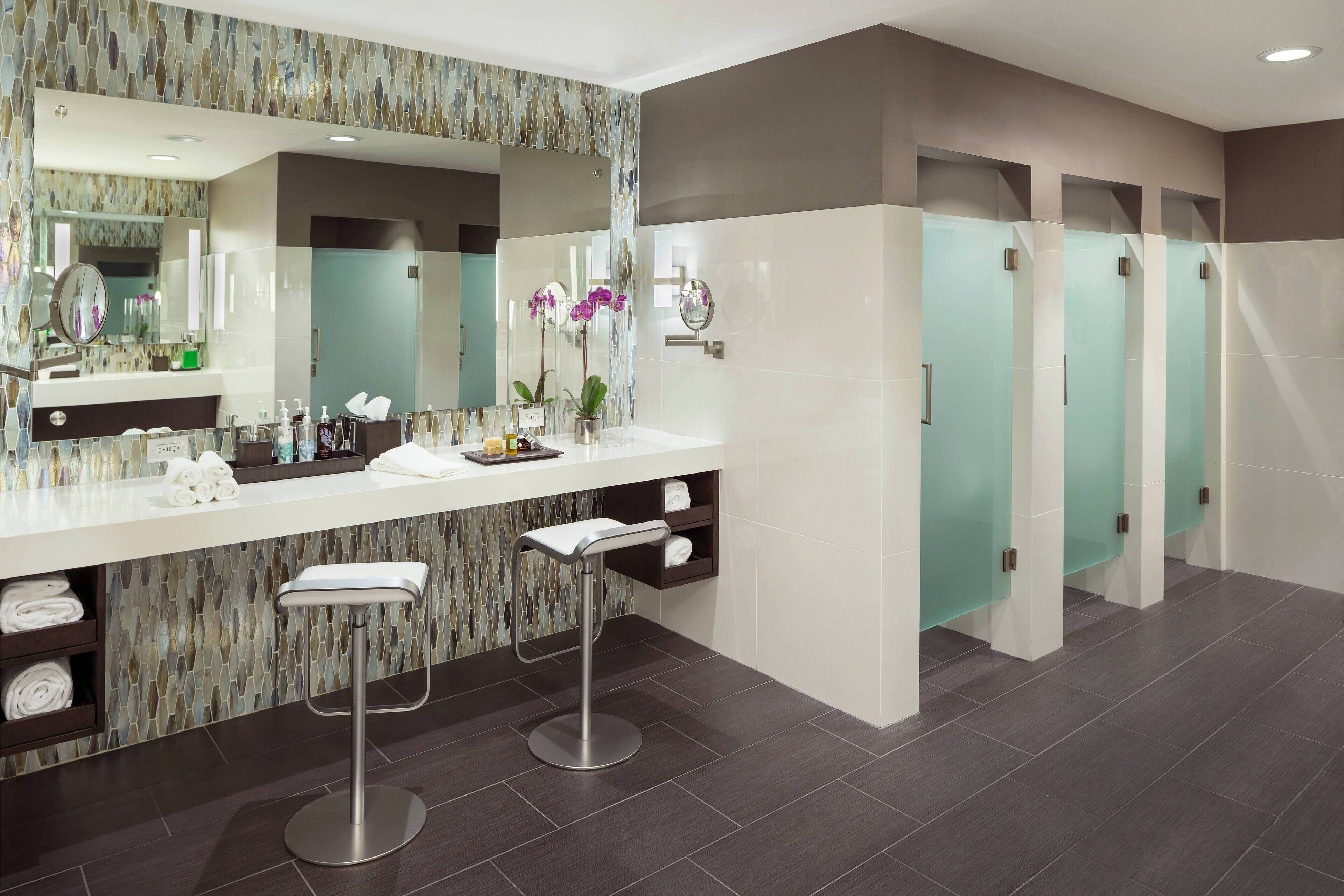 Une salle de bain moderne avec un grand miroir, un meuble-lavabo avec des articles de toilette, un tabouret de bar et une rangée de toilettes séparées par des cloisons en verre.