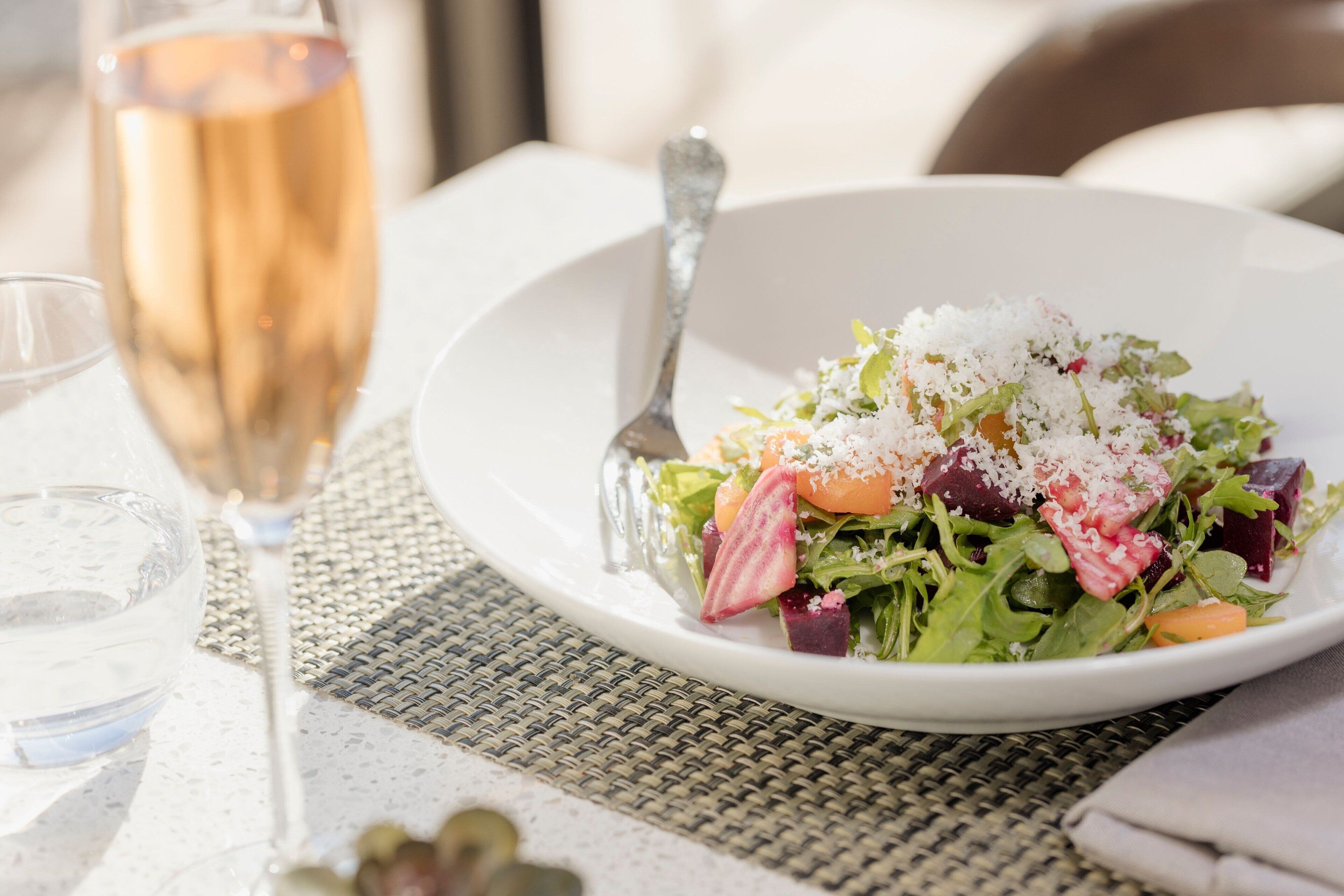 Une assiette blanche contenant une salade, un verre de vin et une fourchette, le tout posé sur une table recouverte d’un set de table en fibres tressées.